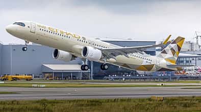 Ein Passagierflugzeug der Fluggesellschaft Etihad Airways startet von einer Landebahn, im Hintergrund sind Industriegebäude zu sehen.