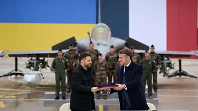 Der französische und ukrainische Präsident schütteln sich die Hände. Im Hintergrund ist ein Rafale-Kampfjet zu sehen.