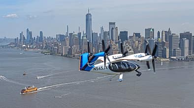 Das Foto zeigt das elektrische Lufttaxi von Joby Aviation im Flug über New York. Das eVTOL mit mehreren schwenkbaren Propellern trägt die Kennung N545JX und fliegt über dem Hudson River; im Hintergrund ist die Skyline von Manhattan mit dem One World Trade Center sichtbar. Laut Dateiname stammt die Aufnahme aus dem Jahr 2026.