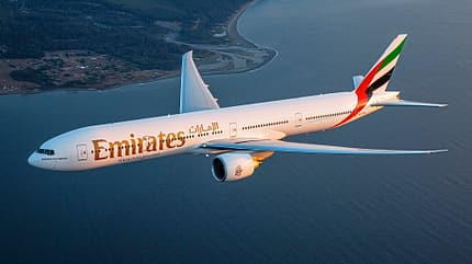 Boeing 777-300ER von Emirates bei einem Flug im Sonnenuntergang über Wasser