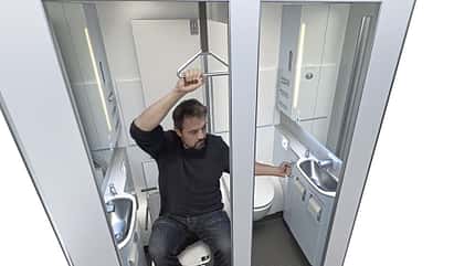 Ein Mann sitzt in einer sehr kleinen, modernen Zug- oder Flugzeugtoilette und hält sich an einer Haltestange fest.