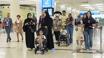 Menschen gehen durch einen Flughafen, darunter Familien mit Kindern, einige im Rollstuhl oder Kinderwagen, sowie Flughafenpersonal.