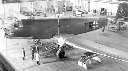 Deutsches Militärflugzeug aus dem Zweiten Weltkrieg in einer Wartungshalle, mehrere Personen arbeiten am Flugzeug.