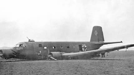 Ein beschädigtes zweimotoriges Militärflugzeug mit deutschen Hoheitszeichen aus dem Zweiten Weltkrieg auf einer Wiese.