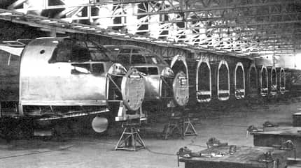 Fertigungslinie mit mehreren im Bau befindlichen Flugzeugrümpfen in einer Industriehalle. Die Aufnahme ist schwarz-weiß und zeigt die Struktur und Montage von Flugzeugteilen.