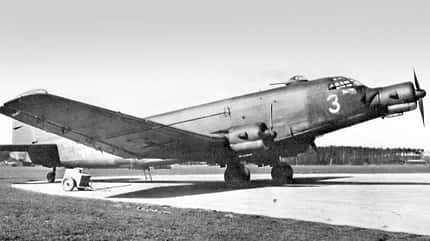 Ein großes, viermotoriges Propellerflugzeug aus der Zeit des Zweiten Weltkriegs steht auf einer Betonfläche eines Flugplatzes. Das Flugzeug hat eine markante, verglaste Bugsektion und Tarnlackierung.