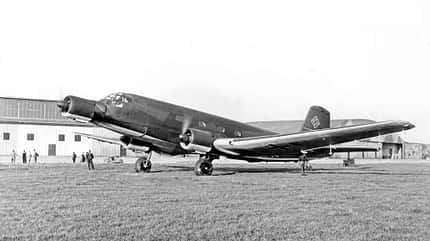Schwarzweiß-Fotografie eines großen viermotorigen Propellerflugzeugs aus der Zeit des Zweiten Weltkriegs auf einem Flugplatz mit Hangar im Hintergrund. Das Flugzeug trägt ein Hakenkreuz auf dem Seitenleitwerk. Mehrere Personen stehen um das Flugzeug herum.