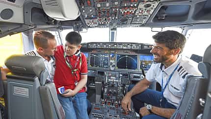 Das Bild zeigt das Cockpit eines Flugzeugs mit zwei Piloten und einem Kind, das auf dem Schoß eines Erwachsenen sitzt. Alle Personen wirken fröhlich und unterhalten sich.