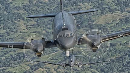 AC-47T, Formationsflug, Turboprop, Propellerflugzeug, Zweimotoriges Militärflugzeug, Frontansicht, Luft-Luft-Aufnahme, Gunship, Militärluftfahrt, 2026 Die Aufnahme zeigt zwei AC-47T im Formationsflug aus frontaler Perspektive über einer grünen Landschaft; das vordere Flugzeug füllt den Bildkader, das zweite folgt in Linie dahinter. Sichtbar sind die laufenden Propellerkreise der zweimotorigen Turboprop-Konfiguration, eine graue Militärlackierung und ein Sensorpod unter dem Bug. Es handelt sich um eine Luft‑Luft‑Aufnahme mit deutlicher Betonung der Nase und Tragflächen des vorderen Musters.
