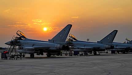 Pitch Black 2026, Luftwaffe, Eurofighter Typhoon, Mehrzweckkampfflugzeug, Militärflugzeug, Militärübung, Bundeswehr, Flugplatzvorfeld, Bodenunterstützung, Sonnenuntergang Das Foto zeigt mehrere Eurofighter Typhoon der Luftwaffe während der Übung Pitch Black. Die Kampfflugzeuge stehen mit geöffneten Cockpithauben auf dem Vorfeld, umgeben von Leitern und Bodenunterstützungsgeräten; die Eisernen‑Kreuz‑Kennzeichen und deutsche Flaggen an den Seitenleitwerken sind sichtbar. Die tief stehende Sonne erzeugt Silhouetten vor einem orangefarbenen Himmel.