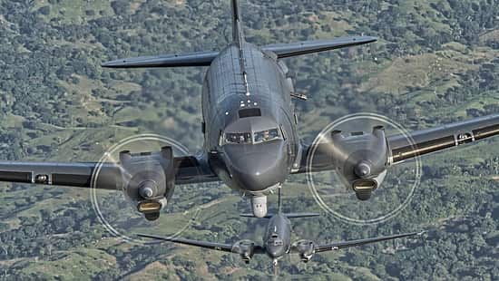 Die Aufnahme zeigt zwei AC-47T im Formationsflug aus frontaler Perspektive über einer grünen Landschaft; das vordere Flugzeug füllt den Bildkader, das zweite folgt in Linie dahinter. Sichtbar sind die laufenden Propellerkreise der zweimotorigen Turboprop-Konfiguration, eine graue Militärlackierung und ein Sensorpod unter dem Bug. Es handelt sich um eine Luft‑Luft‑Aufnahme mit deutlicher Betonung der Nase und Tragflächen des vorderen Musters.
