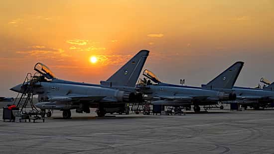 Das Foto zeigt mehrere Eurofighter Typhoon der Luftwaffe während der Übung Pitch Black. Die Kampfflugzeuge stehen mit geöffneten Cockpithauben auf dem Vorfeld, umgeben von Leitern und Bodenunterstützungsgeräten; die Eisernen‑Kreuz‑Kennzeichen und deutsche Flaggen an den Seitenleitwerken sind sichtbar. Die tief stehende Sonne erzeugt Silhouetten vor einem orangefarbenen Himmel.