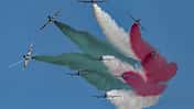 Mehrere Kunstflugzeuge fliegen in Formation und erzeugen farbige Rauchspuren in Grün, Weiß und Rot am Himmel.