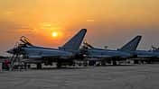 Das Foto zeigt mehrere Eurofighter Typhoon der Luftwaffe während der Übung Pitch Black. Die Kampfflugzeuge stehen mit geöffneten Cockpithauben auf dem Vorfeld, umgeben von Leitern und Bodenunterstützungsgeräten; die Eisernen‑Kreuz‑Kennzeichen und deutsche Flaggen an den Seitenleitwerken sind sichtbar. Die tief stehende Sonne erzeugt Silhouetten vor einem orangefarbenen Himmel.