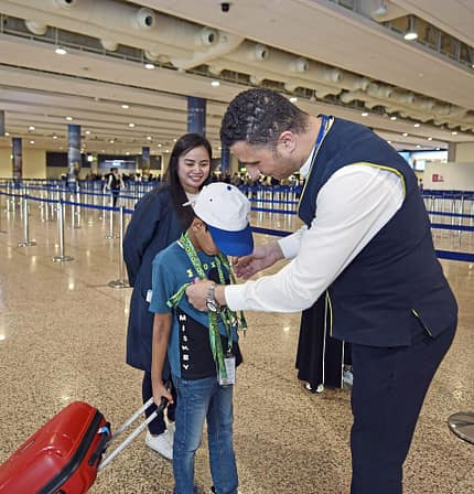 Ein Flughafenmitarbeiter hilft einem Kind mit einem Namensschild, während eine Frau im Hintergrund lächelt. Das Kind zieht einen roten Rollkoffer hinter sich her. Die Szene spielt sich in einer großen, modernen Flughafenhalle ab.