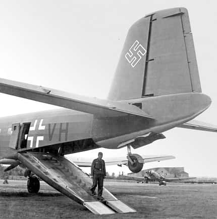 Schwarzweißfoto eines Militärtransportflugzeugs mit geöffneter Heckrampe, ein Soldat steigt aus. Deutlich sichtbare Hoheitszeichen des Dritten Reichs am Leitwerk und Rumpf. Im Hintergrund weitere Flugzeuge und ein Gebäude auf einem Flugfeld.